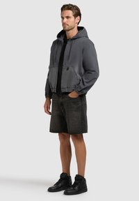 Homme portant une veste à capuche gris foncé, un t-shirt noir, un short en denim noir et des baskets montantes noires, debout avec une main dans la poche.