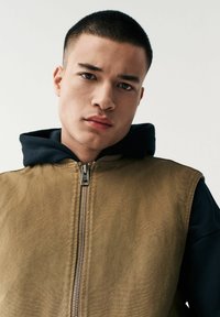 Bruine zip-up vest met een matte afwerking, met daaronder een navy laag met capuchon. Glad stofoppervlak, met een metallic ritsdetail.