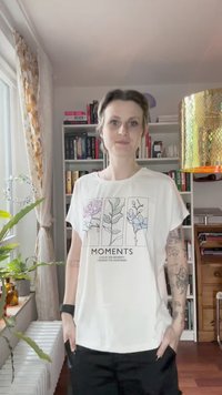 Bela kratka majica s kratkimi rokavi, ki prikazuje cvetlične linijske risbe v pastelnih barvah in besedo "MOMENTS" natisnjeno spodaj. Gladek material, sproščen kroj.