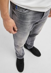 Grå denim jeans med en falmet tekstur, der har et blåt logo-tag på lommen, parret med sorte sneakers.