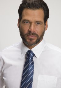 Weißes Hemd mit Brusttasche, kombiniert mit einer dunkelblauen Krawatte mit diagonalen weißen und hellblauen Streifen. Natürlicher Bart und Haare.