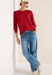Maglione a righe rosse con maniche lunghe abbinato a jeans a gamba larga blu e sneakers beige; la modella è appoggiata a uno sfondo chiaro.