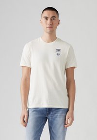Cream-colored short-sleeve cotton t-shirt with a round neck, featuring a blue logo and graphics on the chest. Worn with light blue jeans.