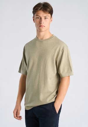Jonge man met kort bruin haar draagt een licht olijfgroen shirt met korte mouwen en donkere broek, staand tegen een effen lichte achtergrond.