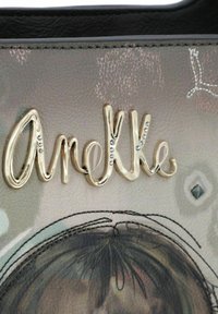Złotą metalową literą „Arielle” zdobioną cyrkoniami na teksturowanym pastelowym tle z portretem dziewczynki i akcentami geometrycznymi.