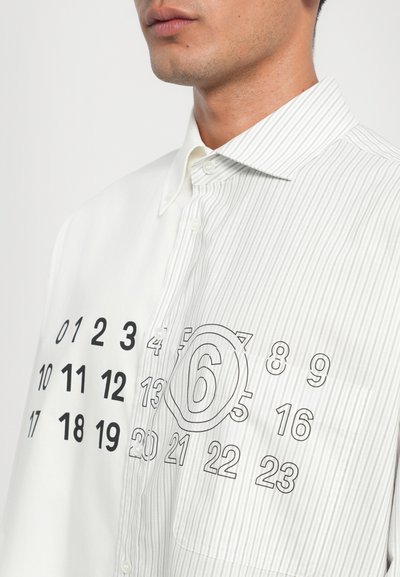 Weiß und grau gestreifte Button-up-Bluse mit großen schwarzen Zahlen, die auf der Vorderseite aufgedruckt sind, und einem Kreis um die Zahl sechs.