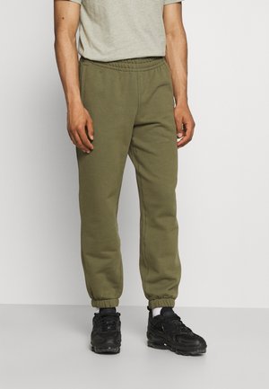 Homme portant un pantalon de jogging vert olive, des baskets noires, des chaussettes blanches et un t-shirt gris clair à manches courtes, debout devant un fond uni.