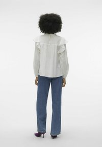 Vit blus med volangärmar, texturerad tyg, detalj med nyckelhål i ryggen; kombinerad med blå vida jeans och lila klackskor.