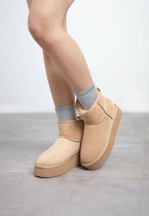 Stivali alla caviglia beige in suede con superficie testurizzata, dotati di una suola spessa e piatta e di un colletto alto e aderente. Indossati con calzini grigi.