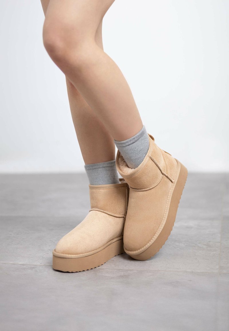 Botas de tobillo beige de ante con una superficie texturizada, que cuentan con una suela gruesa y plana, y un cuello alto y ajustado. Llevadas con calcetines grises.
