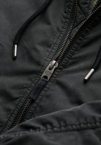 Sort jakke med metallisk lynlåsdetalje, tekstureret stof og en justerbar hætte med sort snor. Inkluderer sølvfarvede hardware-accenter.