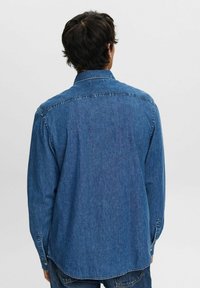 Camisa de mezclilla, azul, de manga larga, con cuello abotonado, vista posterior. Presenta un dobladillo curvado, costuras resistentes y puños con botones.