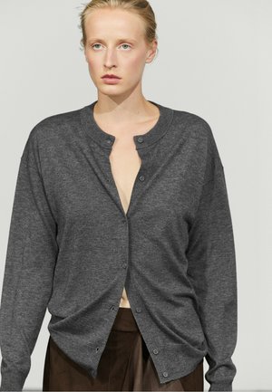 Blonde vrouw met een lichte huid draagt een donkergrijs gebreid vest met knopen over een bruine rok, staand tegen een effen lichte achtergrond.