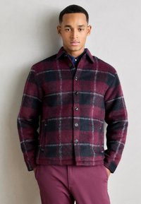 Veste en laine à carreaux dans des nuances de bordeaux, bleu marine et gris, avec un devant boutonné, un col pointu et une texture douce. Modèle associé avec un pantalon assorti.