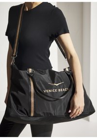 Black nylon bag with gold "VENICE BEACH" logo, featuring short handles and a long strap with gold accents. Smooth texture, spacious design.