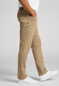 Pantaloni dritti beige con una texture liscia, dotati di una vita con logo, tasche frontali e indossati con sneakers bianche.