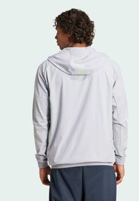 Lichtgrijze sportjas met een capuchon, voorzien van reflecterende neongroene accenten en een elastische zoom, ontworpen voor comfort en beweging.