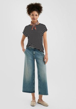 T-shirt à rayures noires et blanches, jean denim large bleu, et chaussures plates en jute beige clair. Une écharpe rouge à motifs nouée autour du cou.