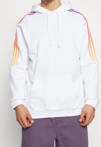 Witte Adidas hoodie met regenboogverloopstrepen op de mouwen, een buidelzak en geborduurd logo, gedragen met paarse broek.