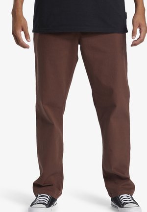 Persona che indossa pantaloni marroni a gamba dritta, maglietta nera e sneakers bianche e nere, in piedi con le braccia rilassate.