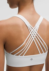 Sujetador deportivo blanco con un diseño de tirantes cruzados en la parte posterior y un logo en la banda inferior. Textura suave con un ajuste ceñido.