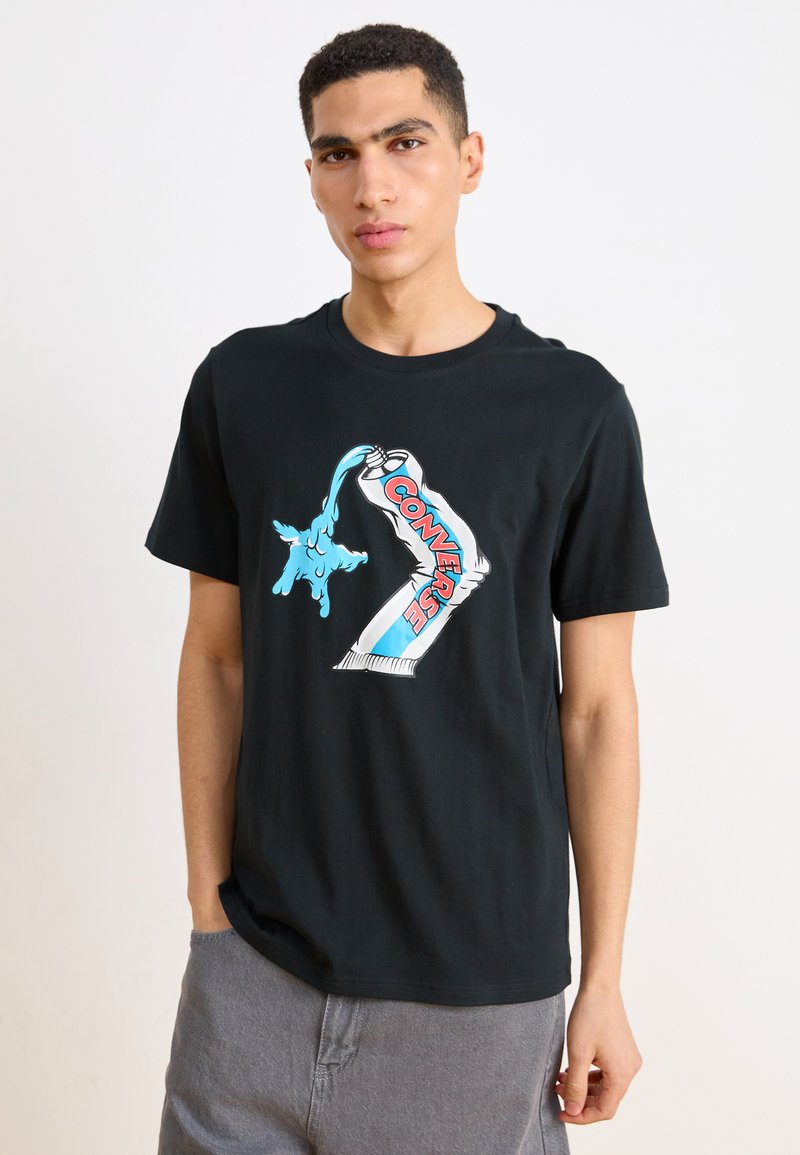 Camiseta de algodón negra con un gran diseño gráfico que presenta acentos en azul y rojo, que representa un logo de Converse y una imaginería juguetona.