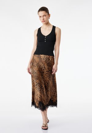 Kobieta ubrana w czarny, bezrękawnik z guzikami oraz midi spódnicę w panterkę z czarną koronkową lamówką, pozująca na białym tle.