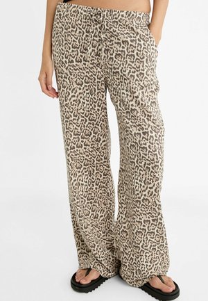 Pantalon ample à imprimé léopard avec taille à cordon, porté avec des tongs à plateforme noires, personne debout avec une main dans la poche.