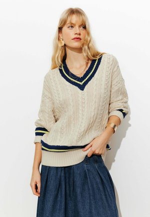 Pull crème en maille torsadée avec col en V, accents rayés bleu marine et vert, et poignets côtelés, porté avec une jupe en jean.