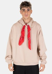 Hoodie oversize beige con tasca a marsupio e un bandana rossa annodata al collo, maniche lunghe e vestibilità rilassata.