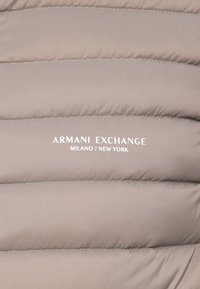 Svijetlosmeđi kviltani materijal s bijelim ispisom "Armani Exchange Milano / New York" u središtu.