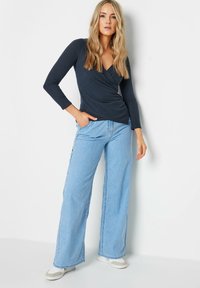 Marineblaue langärmelige Wickelbluse, kombiniert mit hellblauen weiten Jeans. Das Model trägt weiße Sneakers mit beigen Akzenten. Glatte Stoffstruktur.