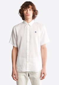 Camisa de lino blanca de manga corta con cuello abotonado y pequeño logo bordado en el pecho. Textura suave y corte holgado.