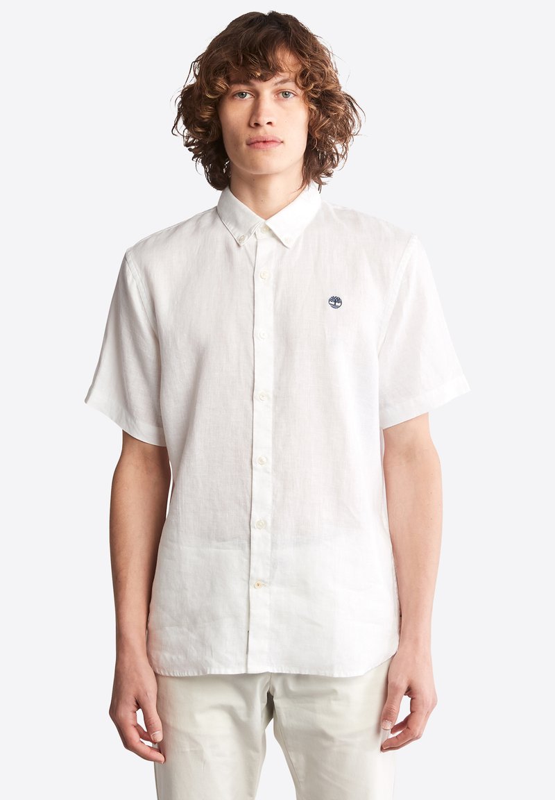 Camisa de lino blanca de manga corta con cuello abotonado y pequeño logo bordado en el pecho. Textura suave y corte holgado.