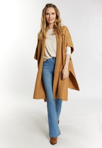 Tan ullkimono med ribbat mönster, bärs över en ljusbeige topp, parat med blå utsvängda jeans och bruna klackstövlar.