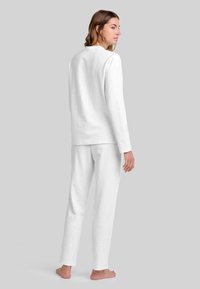 Mujer de pie descalza, vistiendo una camiseta de manga larga blanca sencilla y pantalones blancos a juego, contra un fondo gris.