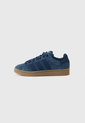 Niedriger Sneaker aus blauem Wildleder mit dunkelblauen Streifen, Schnürsenkeln und Gummisohle auf hellem einfarbigem Hintergrund.