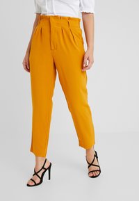 Orange high-waisted trousers with pleats and side pockets, paired with black strappy high heels. Smooth fabric and tapered leg design.