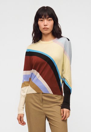 Femme portant un pull à motif abstrait multicolore, un pantalon marron à jambes larges, des chaussures beiges pointues, tenant un sac à main marron.