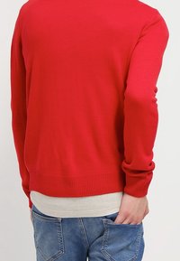 Pull rouge en tricot avec ourlet et poignets côtelés, porté par-dessus une chemise gris clair, associé à un jean en denim bleu.