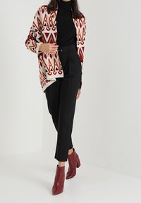 Öppen cardigan med röda, orange och krämfärgade geometriska mönster, ihopparad med svarta skräddade byxor och glansiga bourgognefärgade ankstövlar.