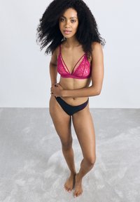 Bralette en dentelle rose avec des bretelles réglables, présentant des motifs floraux complexes. Associé à des culottes noires taille basse, les deux ayant une texture lisse.