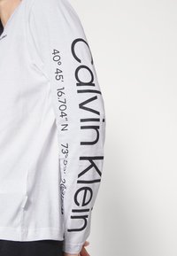 Camisa de manga longa branca com o texto "Calvin Klein" e coordenadas em letras pretas em negrito ao longo da manga esquerda. Tecido com textura suave.