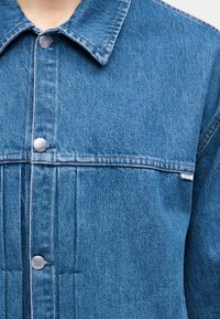 Τζιν μπουφάν με μεταλλικά κουμπιά τύπου κλικ και μια μικρή υφασμάτινη ετικέτα στο στήθος, φορεμένο από ένα άτομο που κόβεται στον λαιμό και τον κορμό.