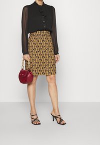 Blouse noire transparente à manches longues avec un col à volants, associée à une jupe longueur genou à motif feuilles or et noir, et des sandales à talons hauts noires.