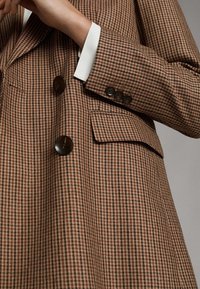 Brązowa marynarka w wzór houndstooth z brązowymi guzikami, klapami w stylu notch i białymi akcentami przy mankietach. Teksturowany materiał o dopasowanym kroju.