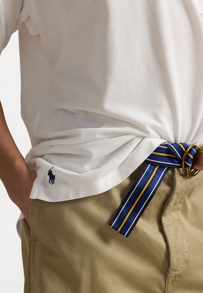 Balts teksturētais polo krekls ar tumši zilu logotipu, kopā ar haki biksēm un zilu, dzeltenu svītrojumu auduma jostu ar brūnu ādas akcentu.