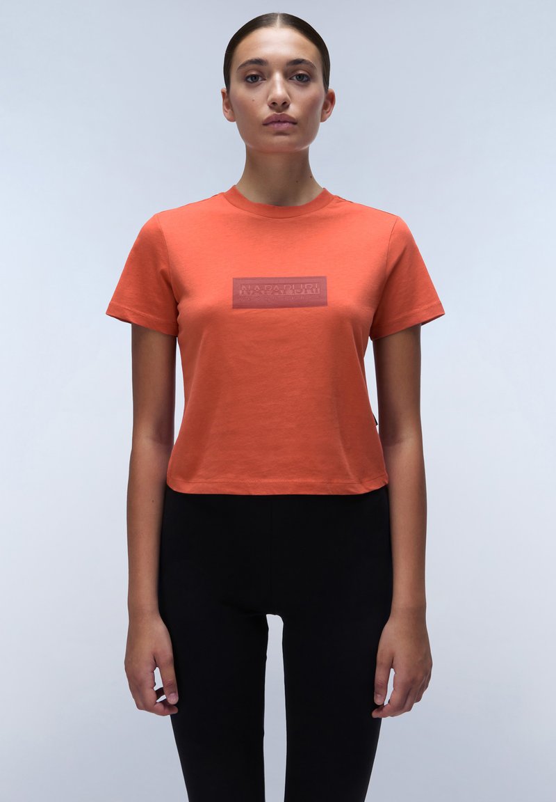 Kurzärmliges, tailliertes T-Shirt aus orangefarbigem Baumwollstoff mit einem rechteckigen, tonalen Logopatch auf der Brust. Getragen mit hoch taillierten schwarzen Leggings.
