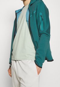 Person med ljusgrön T-shirt, petrolfärgad jacka med dragkedja och dragkedjeficka på ärmen, samt ljusbeiga byxor med händerna i fickorna.