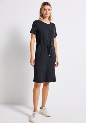 Robe noire à manches courtes avec une coupe décontractée, taille élastique avec cordon de serrage réglable et longueur au genou. Portée avec des baskets blanches.